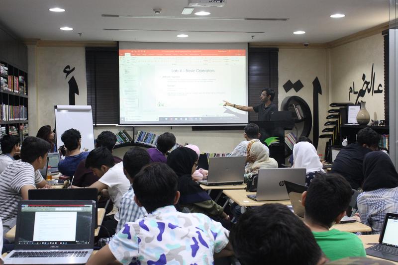 Aazim pointing to a Lab 4 — Basic Operators slide for a full room of students